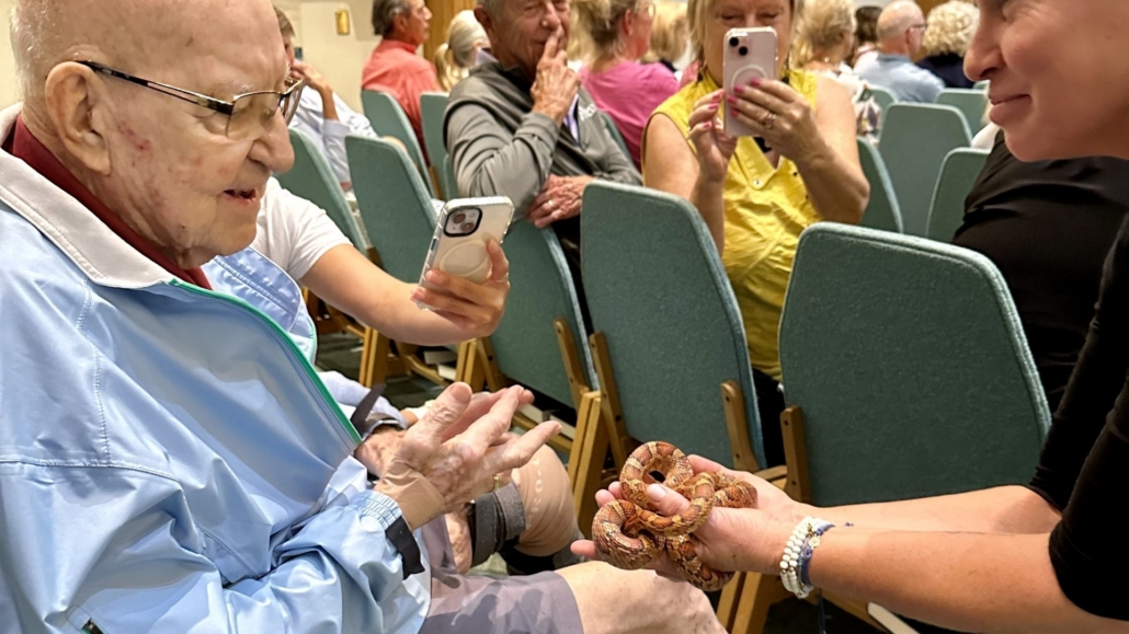 Female speaker smiling as she holds snake for older man to touch while people take photos